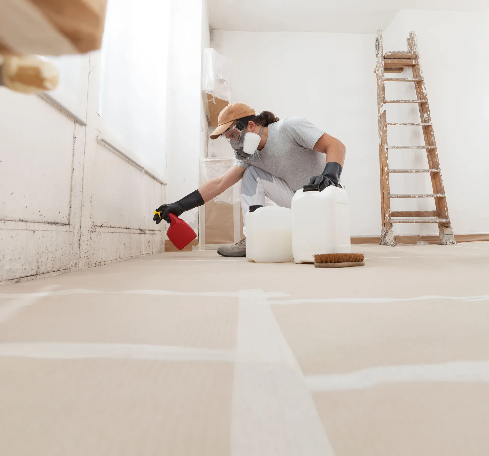 Decorator in a mask and gloves spraying treatment along the base of a wall