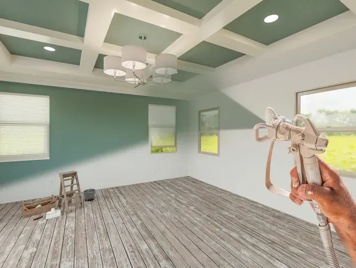 Hand holding an airless spray gun in an empty room with a coffered ceiling